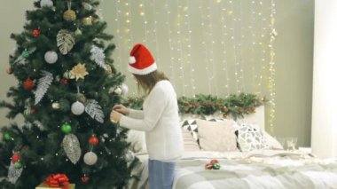 Noel Baba şapkalı genç ve güzel bir kadın Noel ağacını süslüyor. Evinde hediyelerle birlikte yatak odasında takılar takıyor.