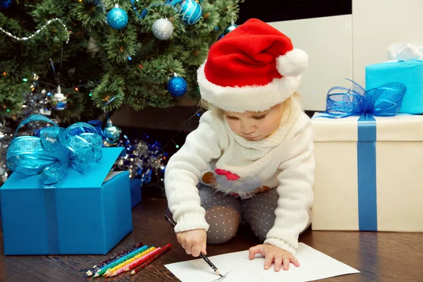 Nettes kleines Mädchen mit Weihnachtsmütze schreibt einen Brief an den Weihnachtsmann in der Nähe des Weihnachtsbaums — Stockfoto