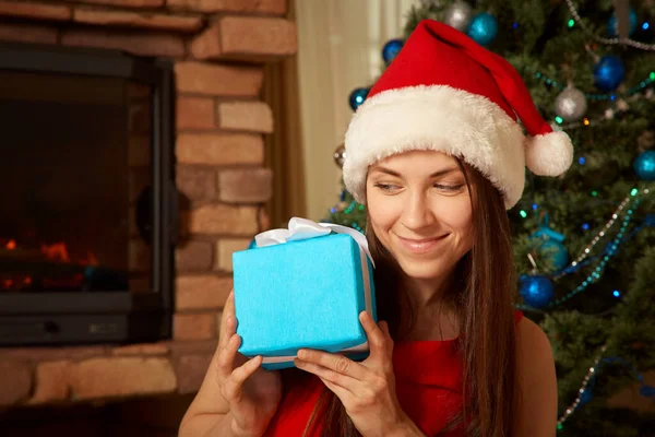 Žena v Santa klobouku s dárkovou krabicí v blízkosti vánočního stromečku. — Stock fotografie