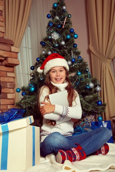 Teenager-Mädchen mit Weihnachtsmütze und Tablet-Computer in der Nähe des Weihnachtsbaums. Weihnachtskind. — Stockfoto
