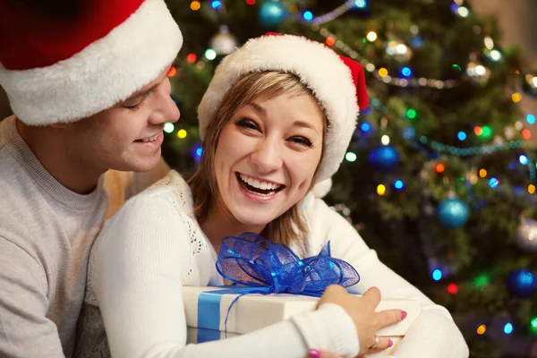 Jeune couple célébrant Noël à la maison. homme et femme en chapeau de Père Noël avec une boîte cadeau — Photo