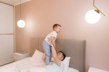 Genç baba, bebeğiyle oynuyor ve gülüyor. Oğlu evde, yatakta, mutlu bir aile.