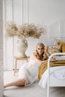 Çekici, kıvırcık saçlı ve makyajlı genç bir kadın. Stüdyonun parlak iç kısımlarında beyaz bir elbise giymiş.