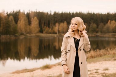 Kıvırcık saçlı ve makyajlı, altın sonbahar gölünün arka planında bej renkli trençkotlu çekici sarışın kadın.