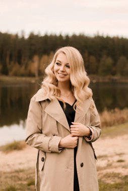 Kıvırcık saçlı ve makyajlı, altın sonbahar gölünün arka planında bej renkli trençkotlu çekici sarışın kadın.