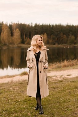 Kıvırcık saçlı ve makyajlı, altın sonbahar gölünün arka planında bej renkli trençkotlu çekici sarışın kadın.
