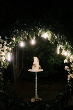 Lüks düğün, çiçeklerle süslenmiş beyaz pasta, arka planda gece çekilmiş fotoğraf.