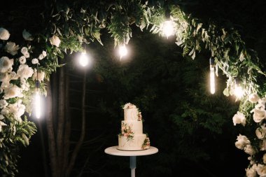 Lüks düğün, çiçeklerle süslenmiş beyaz pasta, arka planda gece çekilmiş fotoğraf.
