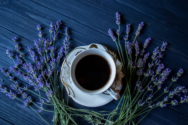 A cup of coffee and lavender flowers on a dark blue wooden background.