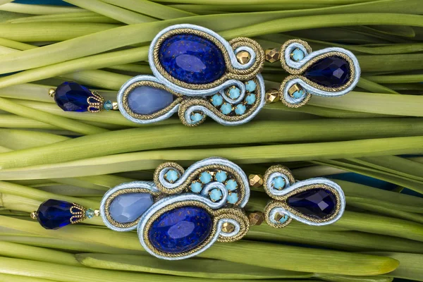 Pendientes azules bijouterie Soutache con piedras azules y cristales de color azul claro y cian sobre el fondo verde de tallos de hierba —  Fotos de Stock