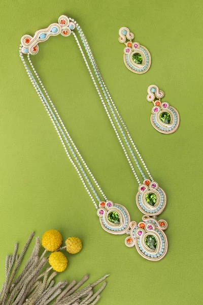 Soutache bijouterie set colorful earrings and necklaces with green purple cyan and pink crystals on green background with grass stems and yellow flowers — Stock Photo, Image