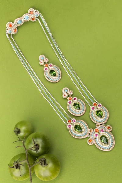 Soutache bijouterie conjunto brincos coloridos e colares com ciano roxo verde e cristais rosa no fundo verde com tomates verdes — Fotografia de Stock