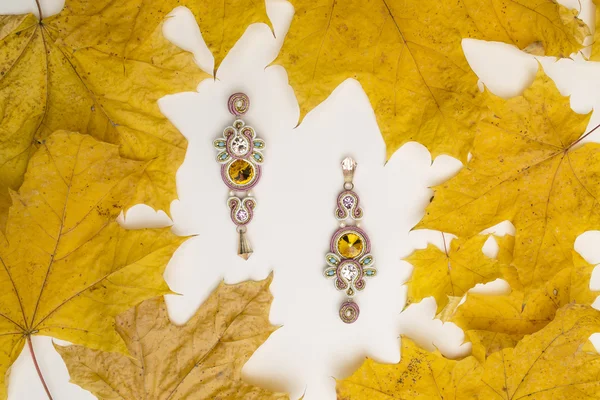 Soutache bijouterie pink earrings with pink yellow and cyan crystals on the white background with yellow maple leaves — Stock Photo, Image