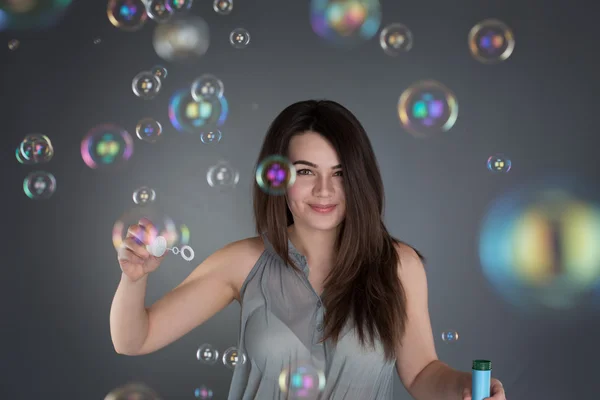 Menina morena jovem inflando bolhas de sabão no fundo cinza Fotos De Bancos De Imagens