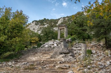 Antik Termessos şehrindeki Hadrian Geçidi 'nin kalıntıları ağaçlarla çevrili ve kayalık bir ön planda.