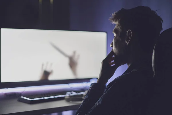 Um jovem homem sentado na frente da tela e assistindo o filme de terror à noite, emoções assustadoras — Fotografia de Stock