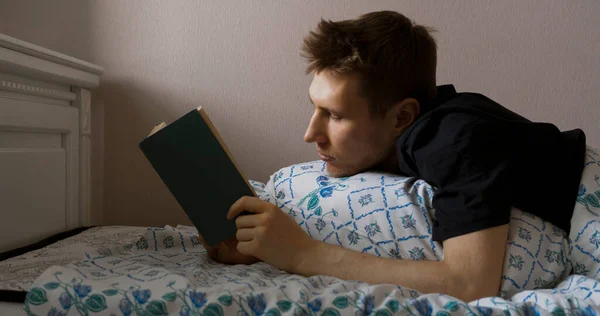 Nuori mies makaa sängyssä ja lukee kirjan illalla ennen nukkumaanmenoa kotona makuuhuoneessa — kuvapankkivalokuva