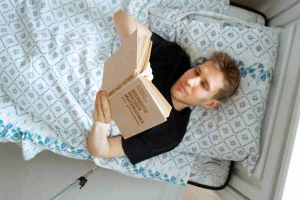 Nuori mies makaa sängyssä ja lukee kirjan illalla ennen nukkumaanmenoa kotona makuuhuoneessa — kuvapankkivalokuva