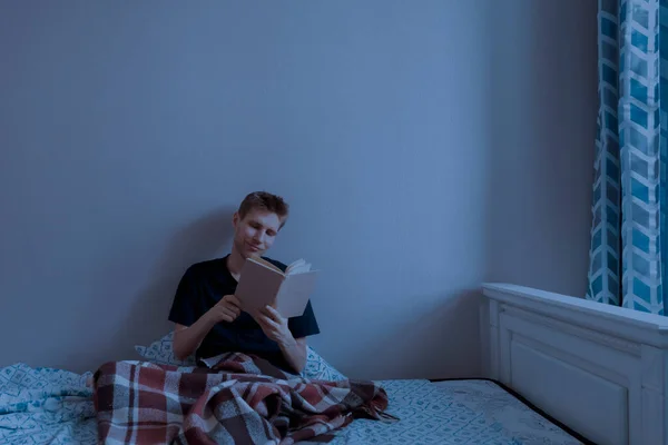 Un giovane maschio sdraiato sul letto e leggere un libro la sera prima di andare a dormire in camera da letto — Foto Stock