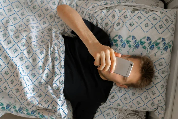 Mies makaa ja käyttää älypuhelinta ennen nukkumaanmenoa, ilta sängyssä verkon riippuvuus — kuvapankkivalokuva