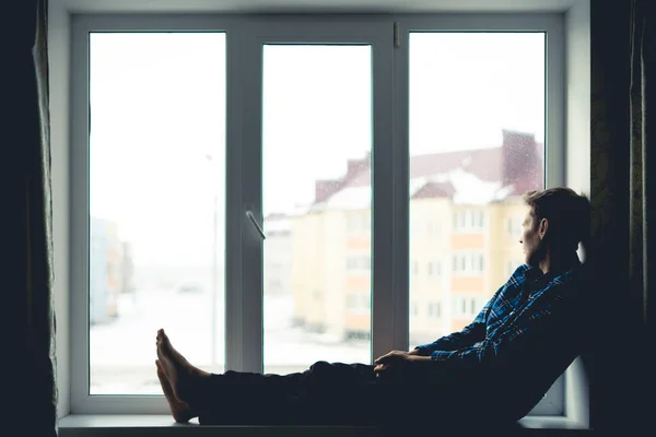 Uma silhueta triste homem solitário pensativo sentado em um peitoril da janela e olhando ao ar livre longe, — Fotografia de Stock