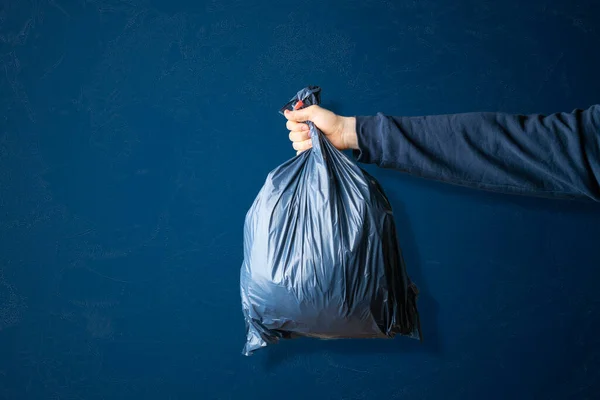 Una mano sosteniendo la bolsa de basura azul oscuro, concepto de limpieza del planeta tierra, aislado contra la pared de color, concepto simple — Foto de Stock