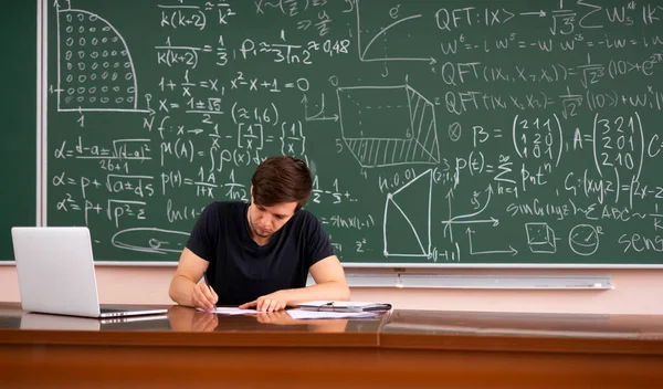 Um jovem professor sentado à mesa perto de quadro-negro e verificar as obras de exame dos alunos — Fotografia de Stock