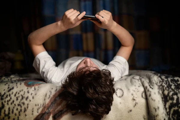 Jovem macho deitado de cabeça para baixo na cama e bate-papo em seu telefone celular gadget f — Fotografia de Stock