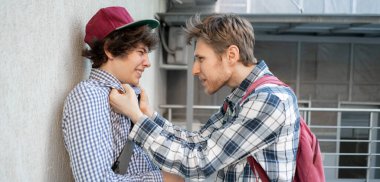 İki adam kavga ediyor, bir yetişkin öğrenciyi tehdit ediyor, duvara yaslanıyor, kabadayılık taslıyor.