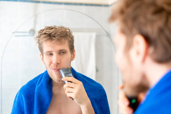 Junger Mann benutzt elektrische Trimmer Rasur gegenüber dem Spiegel b — Stockfoto