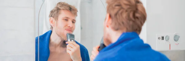 Junger Mann benutzt elektrische Trimmer Rasur gegenüber dem Spiegel b — Stockfoto