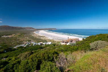Noordhoek Long Beach 'in panorama manzarası. Cape Town yakınlarında beyaz kumlu. Güney Afrika' da mavi gökyüzüne karşı. Chapmans Peak Drive 'da görülmüş. Güzel deniz feneriyle Kommetjie kasabası plajın arka ucunda yer almaktadır.