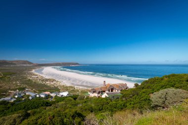 Noordhoek Long Beach 'in panorama manzarası. Cape Town yakınlarında beyaz kumlu. Güney Afrika' da mavi gökyüzüne karşı. Chapmans Peak Drive 'da görülmüş. Güzel deniz feneriyle Kommetjie kasabası plajın arka ucunda yer almaktadır.