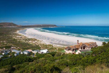 Noordhoek Long Beach 'in panorama manzarası. Cape Town yakınlarında beyaz kumlu. Güney Afrika' da mavi gökyüzüne karşı. Chapmans Peak Drive 'da görülmüş. Güzel deniz feneriyle Kommetjie kasabası plajın arka ucunda yer almaktadır.