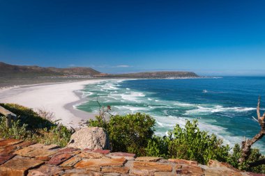 Noordhoek Long Beach 'in panorama manzarası. Cape Town yakınlarında beyaz kumlu. Güney Afrika' da mavi gökyüzüne karşı. Chapmans Peak Drive 'da görülmüş. Güzel deniz feneriyle Kommetjie kasabası plajın arka ucunda yer almaktadır.