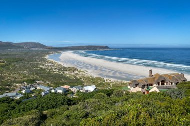 Noordhoek Long Beach 'in panorama manzarası. Cape Town yakınlarında beyaz kumlu. Güney Afrika' da mavi gökyüzüne karşı. Chapmans Peak Drive 'da görülmüş. Güzel deniz feneriyle Kommetjie kasabası plajın arka ucunda yer almaktadır.