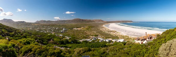 Noordhoek Long Beach 'in panorama manzarası. Cape Town yakınlarında beyaz kumlu. Güney Afrika' da mavi gökyüzüne karşı. Chapmans Peak Drive 'da görülmüş. Güzel deniz feneriyle Kommetjie kasabası plajın arka ucunda yer almaktadır.