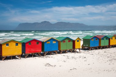 Famous colorful beach houses in Muizenberg near Cape Town, South Africa with mountains of Cape of Good Hope Peninsula in the background against blue sky with clouds