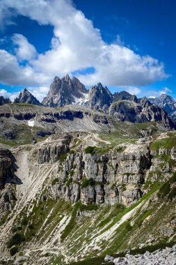 İtalya 'nın Tre Cime bölgesinden görülen Cadini di Misurina dağ grubunun manzara görüntüsü; sivri Dolomite kuleleri, katmanlı uçurumlar ve küçük kar yamaları ile mavi bulutlu gökyüzüne karşı yeşil yamaçlar