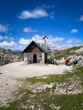 Küçük dağ şapeli Cappella degli Alpini Tre Cime di Lavaredo yürüyüş parkurunda; kayalık bir yaylada taş çan kulesi yürüyüşçüleri ve Dolomite zirveleri bulutlu mavi gökyüzüne karşı