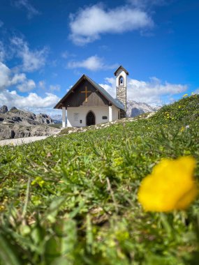Cappella degli Alpini 'nin alçak açılı görüntüsü Tre Cime yürüyüş yolu boyunca bulanık alp çayırı ve sarı bir düğün çiçeği (Ranunculus) ile bulutlu mavi gökyüzüne karşı