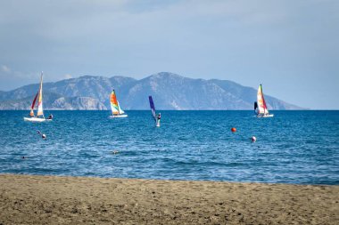 Dalaman yakınlarındaki Sarigerme 'de, renkli yelkenli tekneleri ve sakin Akdeniz sularında, berrak bir gökyüzünün altında bir dağ zeminiyle rüzgar sörfçüsü.