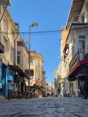 Istanbul, Turkey - October 6, 2025: Low anlgle view of narrow cobblestone Gozlukcu Sokak in Ortakoycafes and small shops with signs and balconies under a clear blue sky.