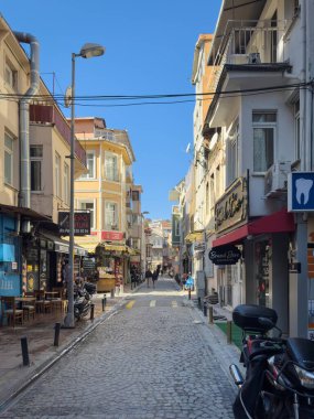 İstanbul, Türkiye - 6 Ekim 2025: Ortakoycafeler ve balkonlu küçük dükkanlar, kaldırım kenarındaki scooterlar ve açık mavi gökyüzü altındaki yayalar.