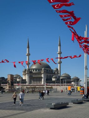İstanbul, Türkiye - 6 Ekim 2025: Taksim Camii, Taksim Meydanı 'nda yürüyen insanlar, çerçevede asılı kırmızı Türk bayrakları ve açık mavi gökyüzüne karşı yükselen ikiz minareler