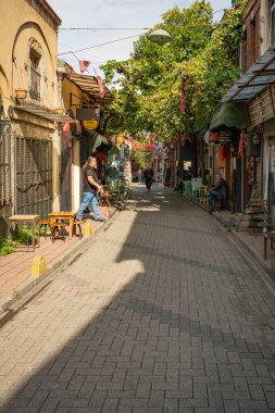 İstanbul, Türkiye - 8 Ekim 2026: Balat 'ta eski binalar, küçük kafeler ve dükkanlar ile güneşli bir ara sokak ve İstanbul, Türkiye' de birkaç yayanın bulunduğu cadde üzerinde bir asma kat gölgesi.