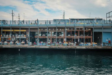 İstanbul, Türkiye - 8 Ekim 2026: Galata Köprüsü altındaki dükkanlar ve restoranlar, yukarıdaki yayalar ve aşağıdaki dalgalı sular, mavi gökyüzü ve bulutlar altında
