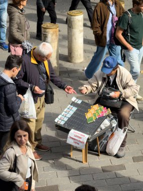 İstanbul, Türkiye - 8 Ekim 2025: Sokak satıcısı Kadıköy, İstanbul, Türkiye 'de kalabalık bir meydanda yoldan geçenlere piyango bileti sattı