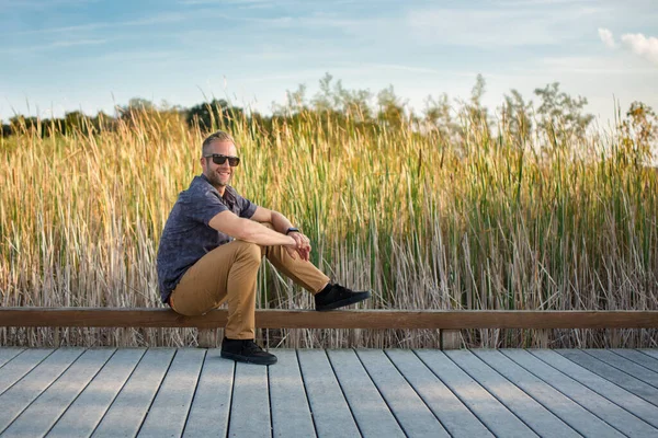 Smiling Man Sunglasses Sits Boardwalk Front Tall Grass — Zdjęcie stockowe