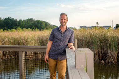 Kendine güvenen, gülümseyen bir adam, bataklık parkındaki korkuluklara yaslanır.